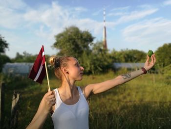 Woman holding flag of latvia and fruit while standing on field against sky