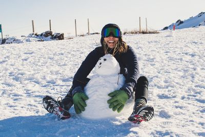 Young woman wearing ski goggles sitting by snowman on field against sky