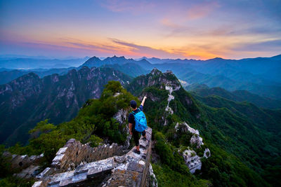 High angle view of male hiker with backpack standing on mountain against cloudy sky during sunset