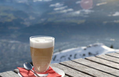 Glass of latte macchiato caffee latte on a wooden table and sunray. snow covered mountain background 