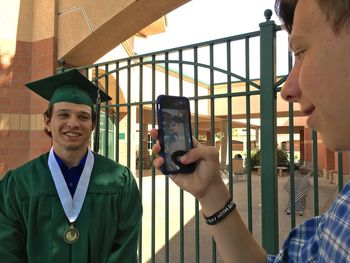 Man photographing brother in graduation gown at college campus