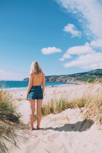 Rear view of young woman standing at beach against sky during sunny day