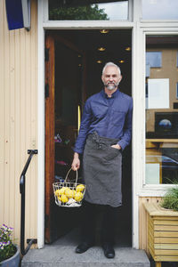 Portrait of confident salesman holding fruit basket while standing at store entrance