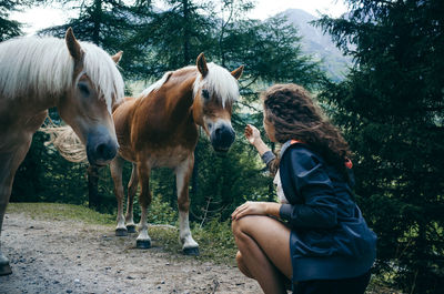 Rear view of woman crouching on field with horses against sky