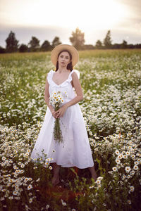 Beautiful young brunette woman in a hat and a white dress standing on a chamomile field at sunset