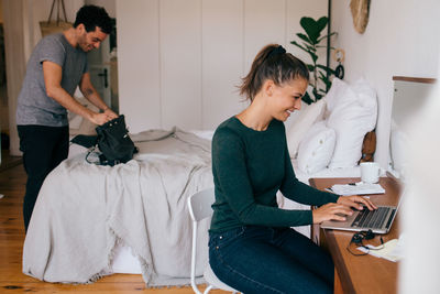 Smiling woman using laptop while boyfriend looking in purse at home