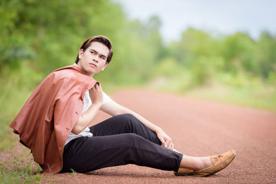 Side view of young man sitting on dirt road against trees