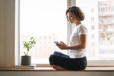 Young brunette woman in casual clothes using mobile phone sitting on window sill at home