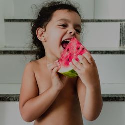 Shirtless happy girl eating watermelon while standing against wall