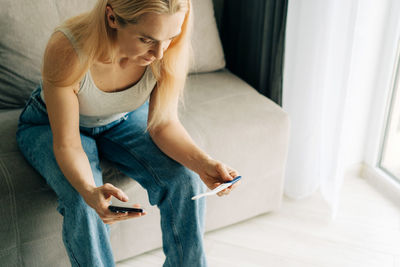 Woman with pregnancy test and mobile phone on sofa at home.