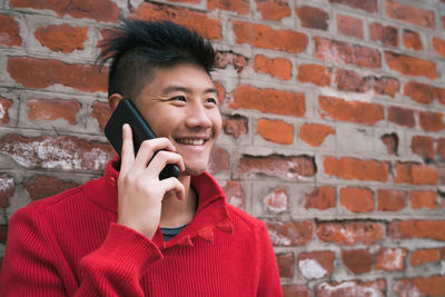 Portrait of smiling young woman using mobile phone against brick wall