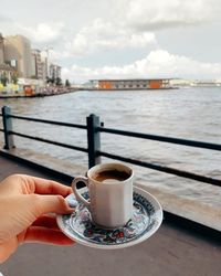 Hand holding coffee cup by railing against sky