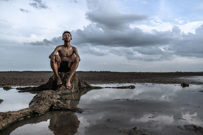 Shirtless boy sitting on rock against cloudy sky during arid climate