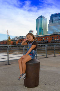 Young woman sitting on shore in city against sky