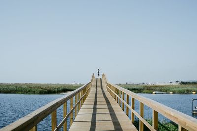 Rear view of young man with backpack standing on footbridge over river against clear sky
