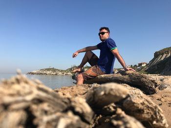 Full length of man sitting on rock at beach against clear blue sky