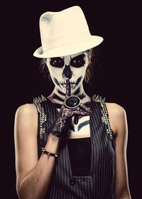 Portrait of woman with skull face paint while finger on lips against black background