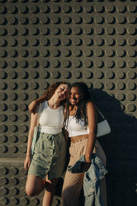 Portrait of happy teenage girl standing with arm around friend against wall during sunny day