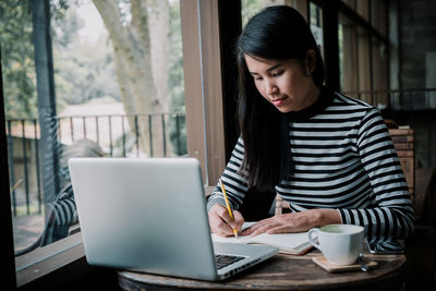 Woman using laptop while writing on book at table in cafe