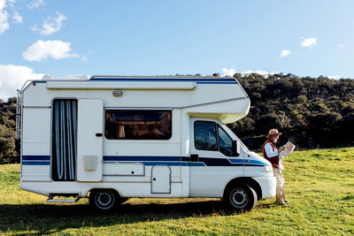 Side view mature male traveler examining paper map while leaning on rv car during road trip in nature on summer day