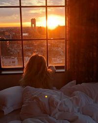 Rear view of woman looking through window while lying on bed at home