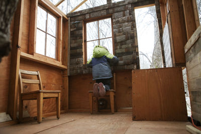 Rear view of boy looking through window while kneeling on chair in tree house