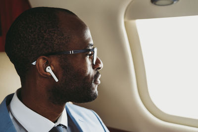 Male entrepreneur with wireless earphones looking through window in private jet