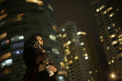 Low angle view of woman wearing hijab standing against illuminated buildings at night