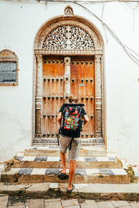 Solo male backpacker with backpack covered in flags stands with ornate door on stone town, zanzibar.