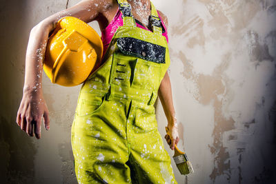 Midsection of woman coveralls holding paintbrush and hardhat against white wall