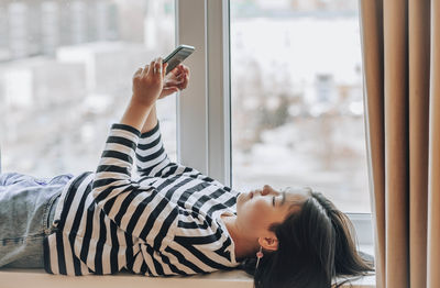 Asian girl relaxes on window with phone, peaceful moments with technology. cityscape outside