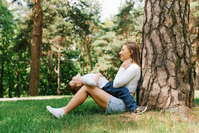 Young caucasian mom and her three-year-old son have fun sitting in the park near a tree on the grass