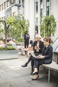 Full length of businesswomen using laptop while sitting on bench with colleagues in background office yard