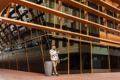 Casual wear man standing against office building while using mobile
