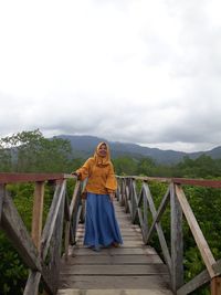 Woman standing on footbridge against sky