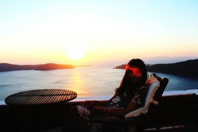 Portrait of young woman sitting on chair at balcony against sky during sunset