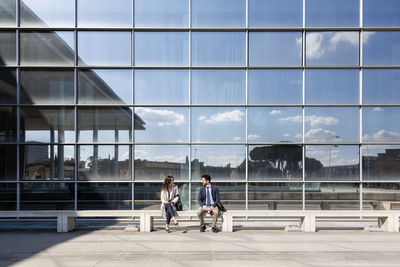 Business couple talking while sitting on bench during sunny day