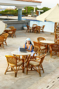 A young woman in a hat at a pool table at a hotel among palm trees drinks a cocktail and works 