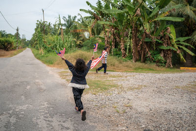 Rear view full length of girl holding malaysian flags while walking on road 