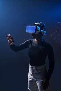 African american female in modern vr headset standing on black background with raised hands while exploring virtual reality in studio