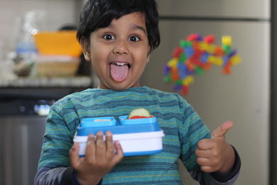 Portrait of boy holding lunch box while sticking out tongue