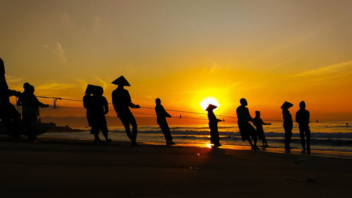 Silhouette people pulling ropes on beach against sky during sunset