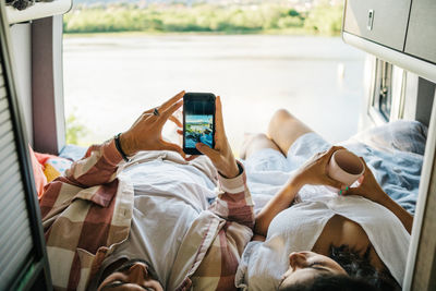 Hispanic couple of travelers lying on bed in camper and taking picture of lake on smartphone during summer trip