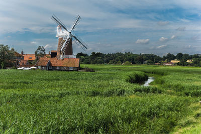 Traditional windmill by grassy field against cloudy sky on sunny day