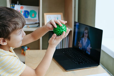 8 years old boy sit by desk with laptop and do exercise with massage ball. smiling woman on screen