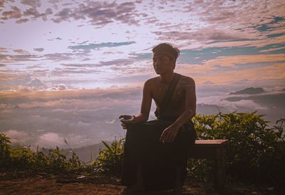 Young man holding drink sitting against sky during sunset