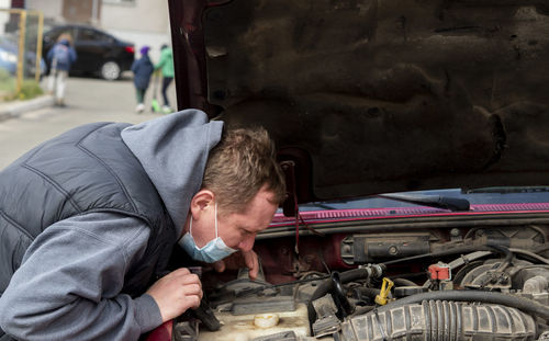 Mechanic in a protective medical mask checks a car engine during a covid-19 coronavirus pandemic