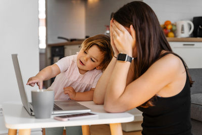 Stressed woman using laptop while daughter pointing at home