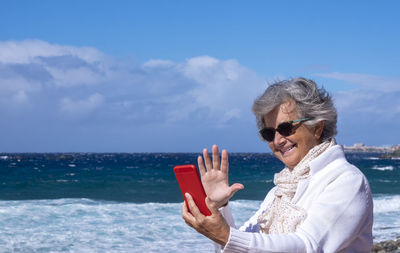 Smiling senior woman talking on video call at beach against sky