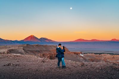 Couple embracing while standing on mountain against clear sky during sunset